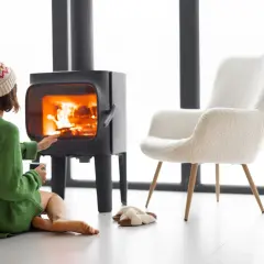 Kobieta w zielonym swetrze i czapce grzeje dłonie przy płonącym kominku, który jest podłączony do komina. Obok stoi puszyste krzesło.