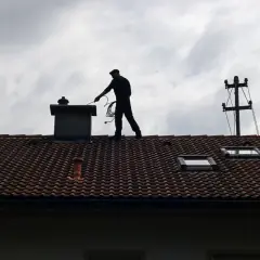 Przegląd kominiarski na dachu. Sylwetka mężczyzny przy kominie, z narzędziami w ręku, na tle pochmurnego nieba.