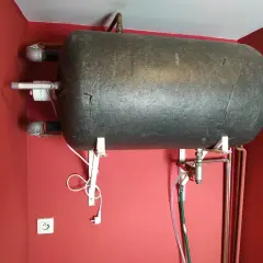 Czarny bojler wiszący na czerwonej ścianie. Widać, że bojler cieknie od spodu, a pod nim biegną rury i wąż.