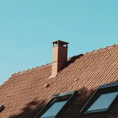 Brązowy, ceglany komin na dachu z dachówkami, pod błękitnym niebem. Może to być mokry komin na strychu po deszczu.