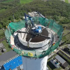 Rozbiórka komina w domu przebiega sprawnie. Koparka pracuje na szczycie, demontując betonową konstrukcję. Zielona siatka zabezpiecza teren.