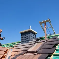 Pracownik na dachu układa dachówki. W tle komin. Zapytaj o szlamowanie komina cena.