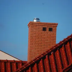 Nowy, ceglany komin z metalowym wkładem i daszkiem, widoczny na tle błękitnego nieba, nad czerwonym dachem.
