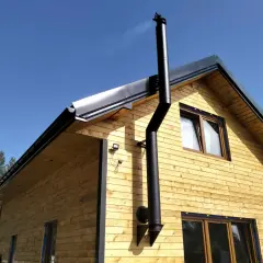Czarny, metalowy komin do kozy w garażu, zamontowany na drewnianej ścianie budynku.