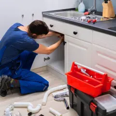 Hydraulik naprawia instalację pod zlewem. Na podłodze części rur i narzędzia. Cennik usług hydraulicznych bez materiału.
