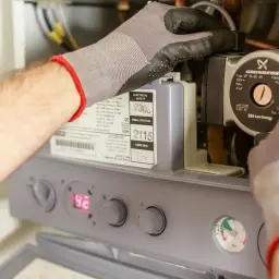 Ręce w rękawiczkach majstrują przy bojlerze, sprawdzając jego elementy. Może to być część instrukcji, jak sprawdzić grzałkę w bojlerze.