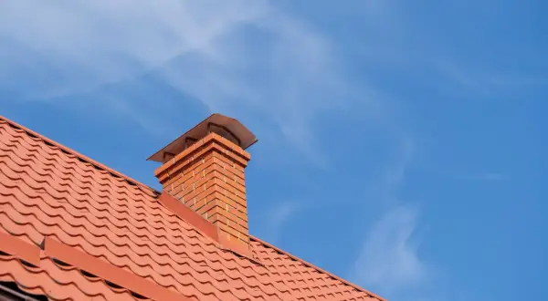 Ceglany komin na dachu pokrytym pomarańczową dachówką. Opierzenie komina wykonane z blachy trapezowej.