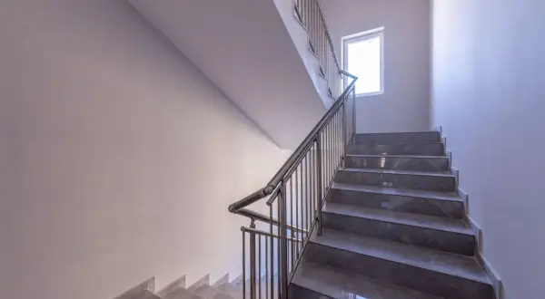 Nowoczesne schody w bloku z szarymi stopniami i metalową balustradą. Jasne ściany i okno zapewniają dobre oświetlenie, tworząc przyjazną atmosferę. Ogrzewanie klatki schodowej w bloku zapewnia komfort.