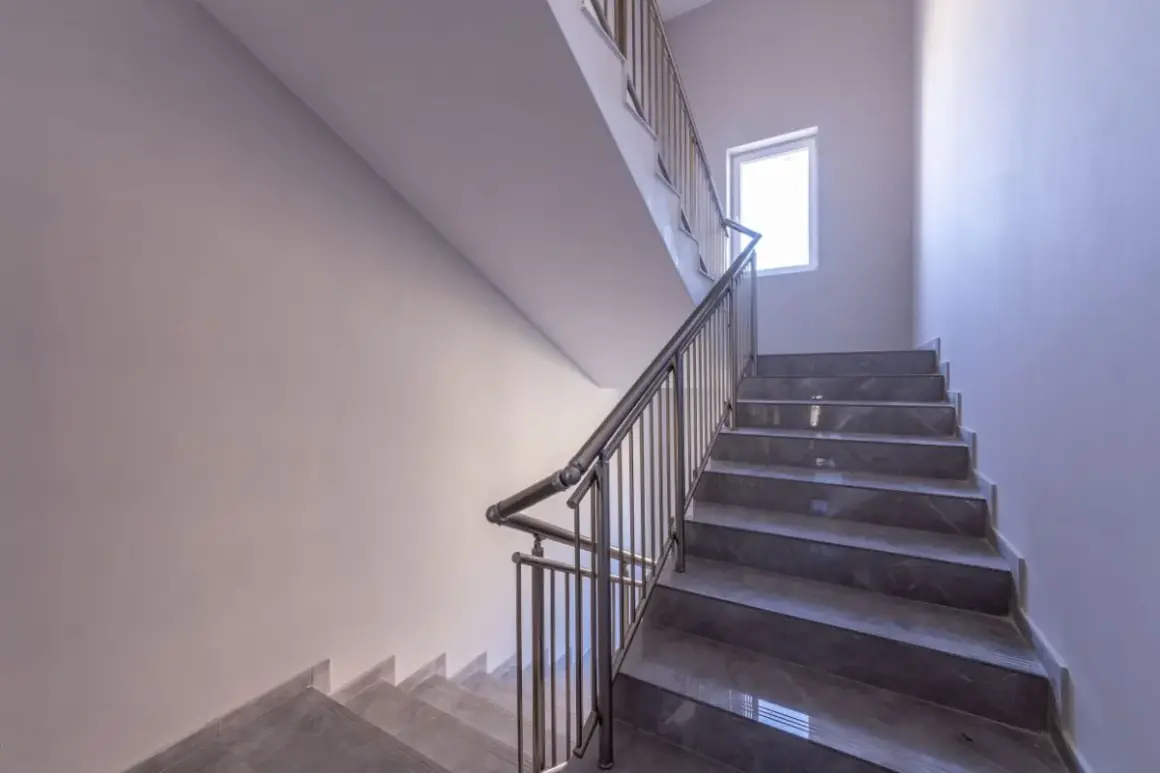 Nowoczesne schody w bloku z szarymi stopniami i metalową balustradą. Jasne ściany i okno zapewniają dobre oświetlenie, tworząc przyjazną atmosferę. Ogrzewanie klatki schodowej w bloku zapewnia komfort.