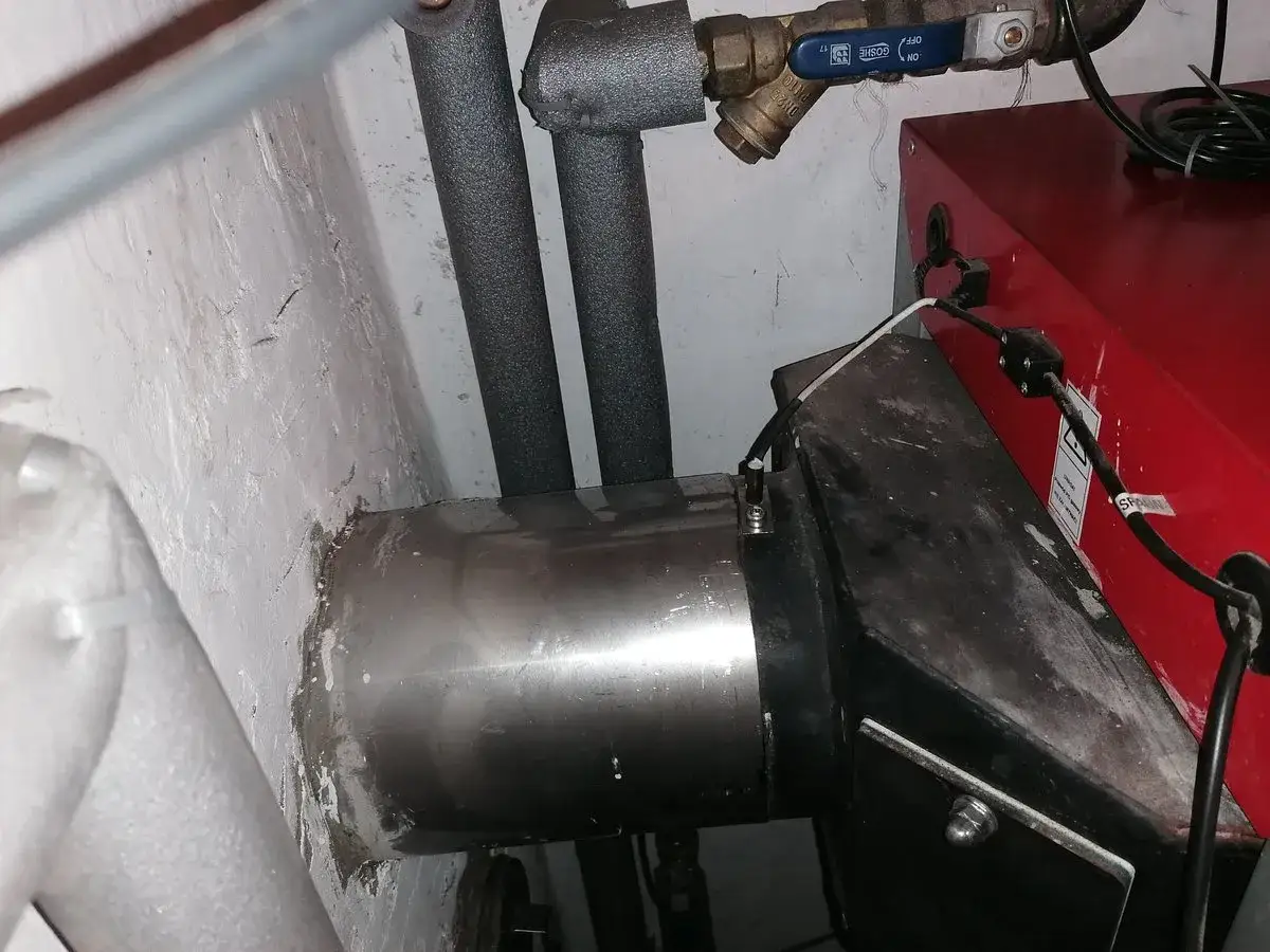 Czujnik spalin do pieca zamontowany na metalowej rurze, blisko czerwonego urządzenia.